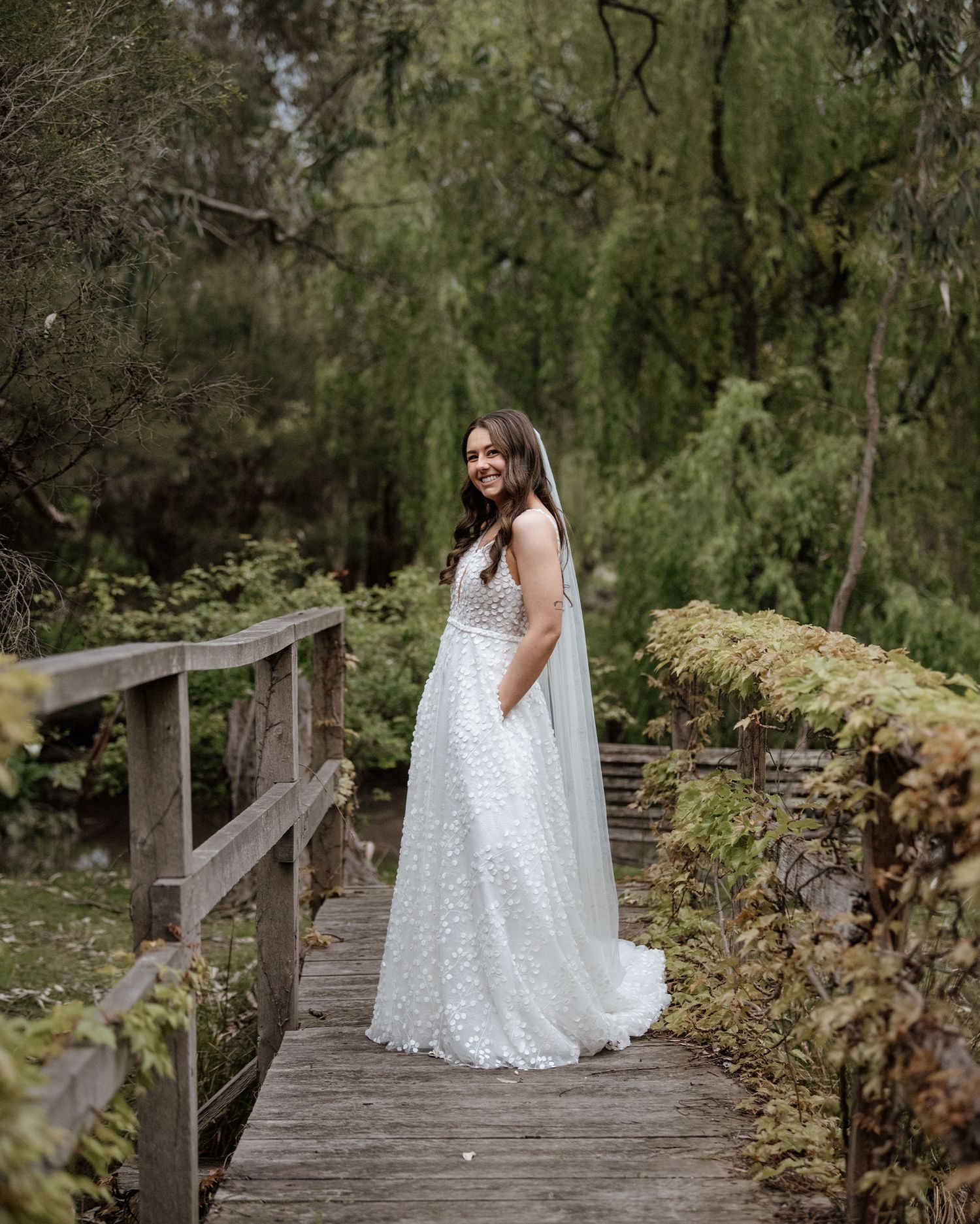 Beautiful wedding dress photo on a rustic wooden bridge surrounded by lush greenery and mossy stone walls.