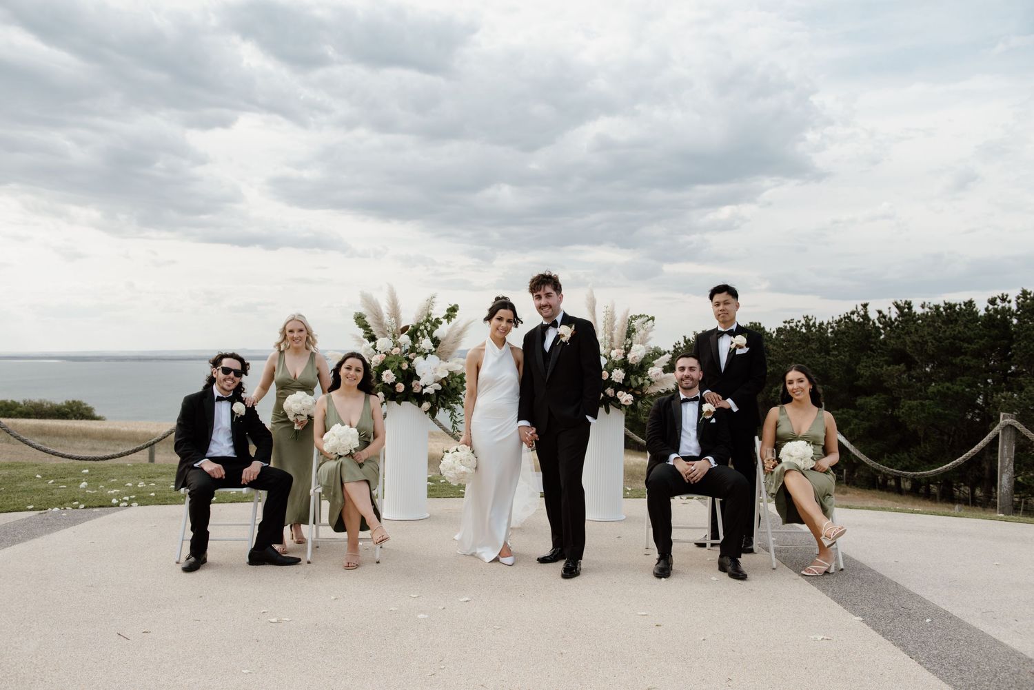 A wedding party poses together at the beach on a cloudy day wearing formal attire and dresses in neutral tones.