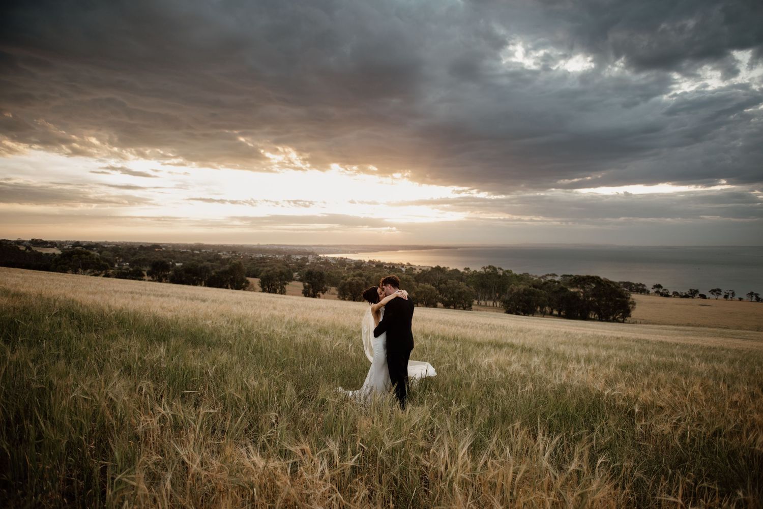A couple in wedding attire stands in a golden grass field at sunset with dramatic clouds overhead.