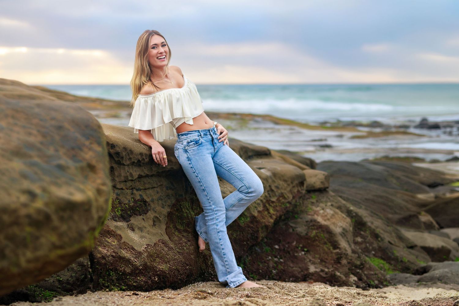 A high school senior in a crochet vest and white cowboy boots poses outdoors in natural sunlight.