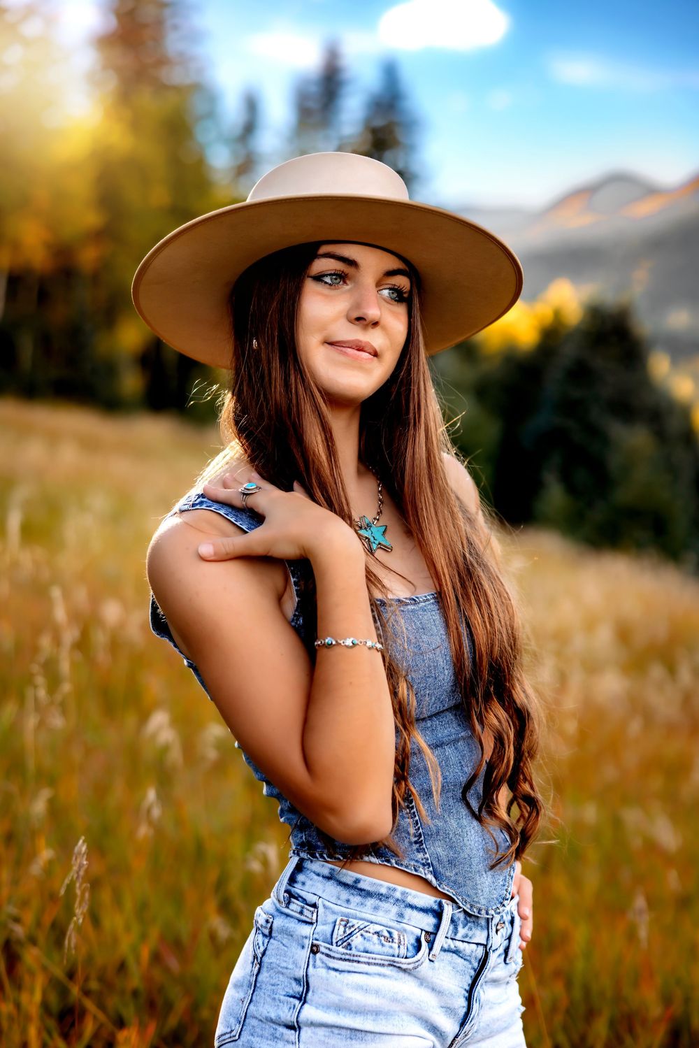 Woman in denim overalls and wide brim hat poses in golden field during sunset with mountains in background. Lake Isabel, Pueblo Photographer