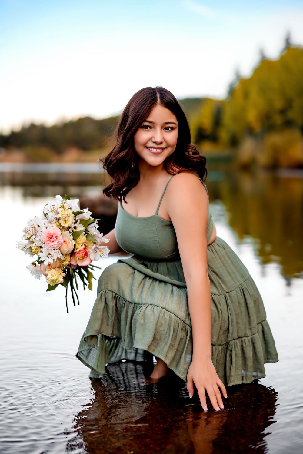 A person in a green dress holds a bouquet of pink and white flowers while posing in a lake during autumn. Senior Pueblo Photographer