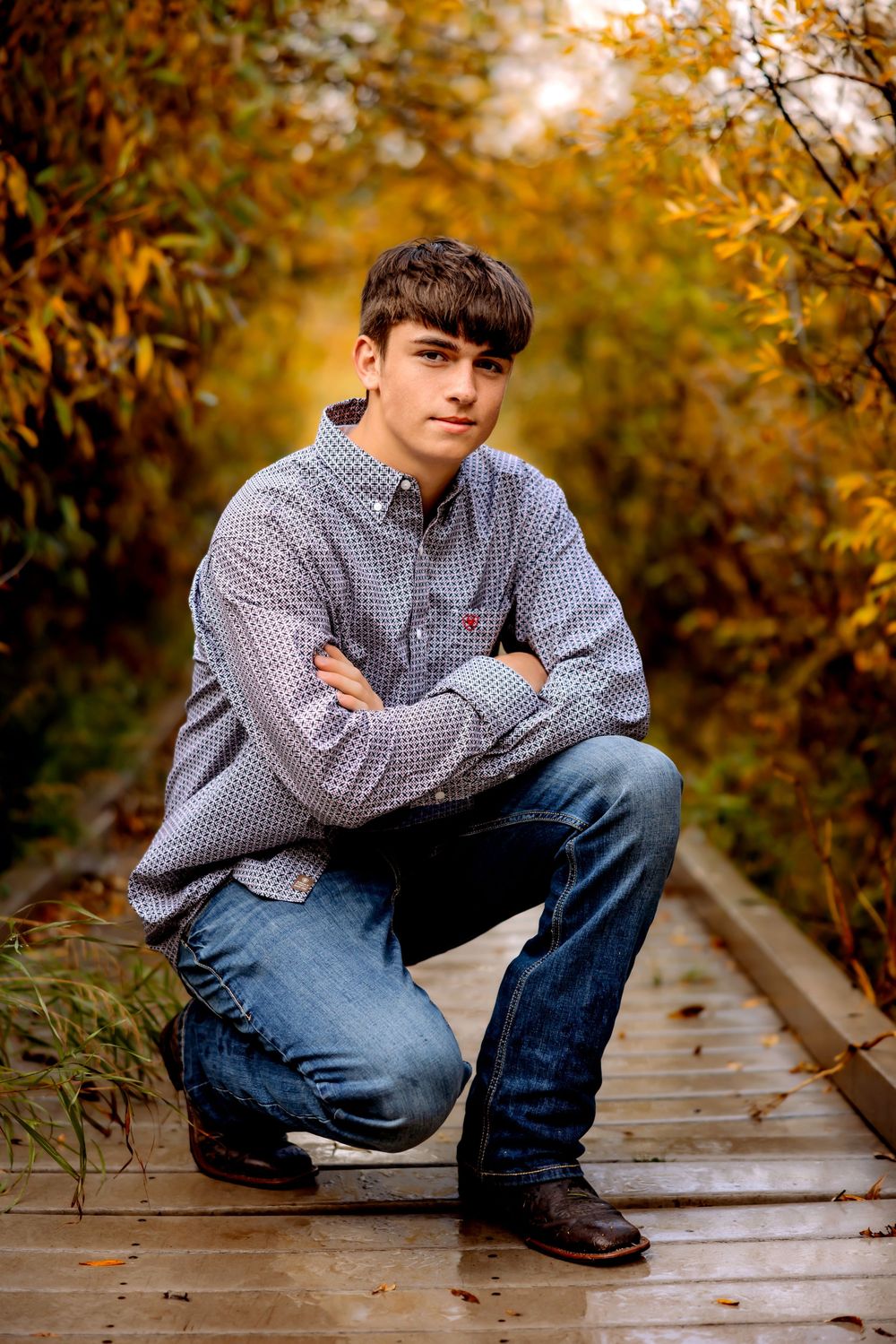 Portrait photo on wooden boardwalk with autumn foliage in background showing person in plaid shirt and jeans.