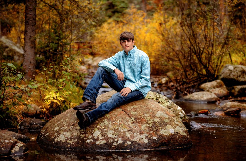 A person in blue sits on a large rock by a tranquil stream surrounded by autumn foliage.