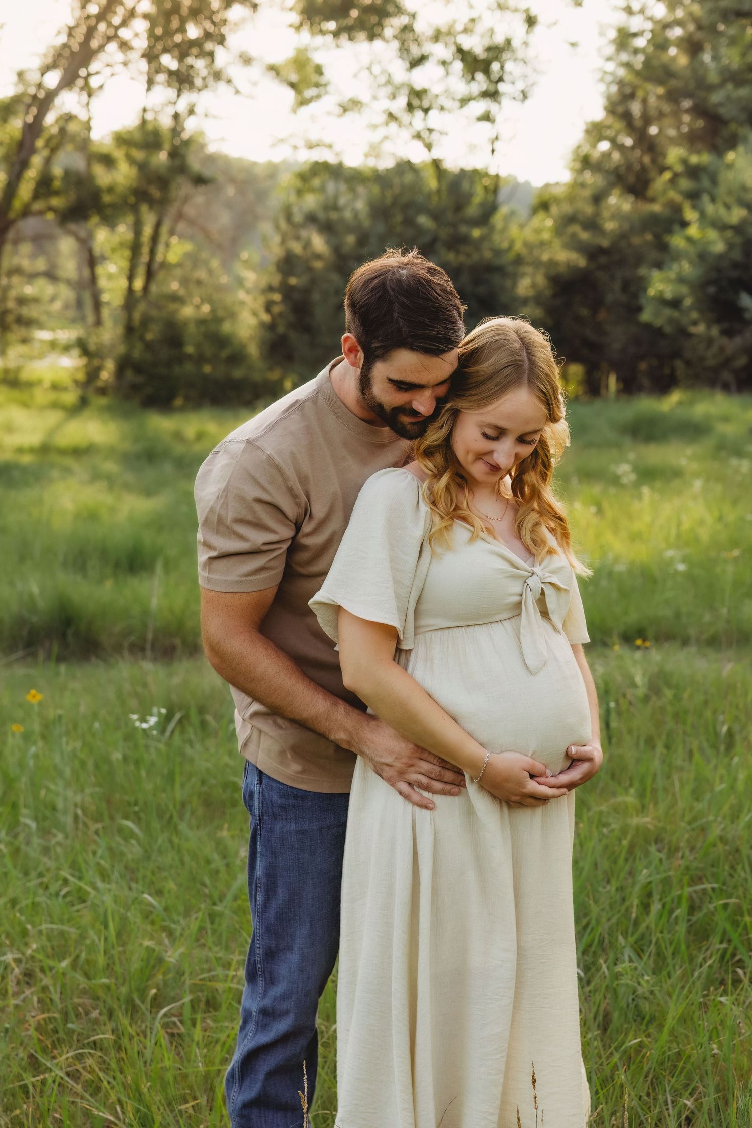 Couple sharing a tender moment outdoors, man embracing pregnant woman in cream dress in a lush green meadow.