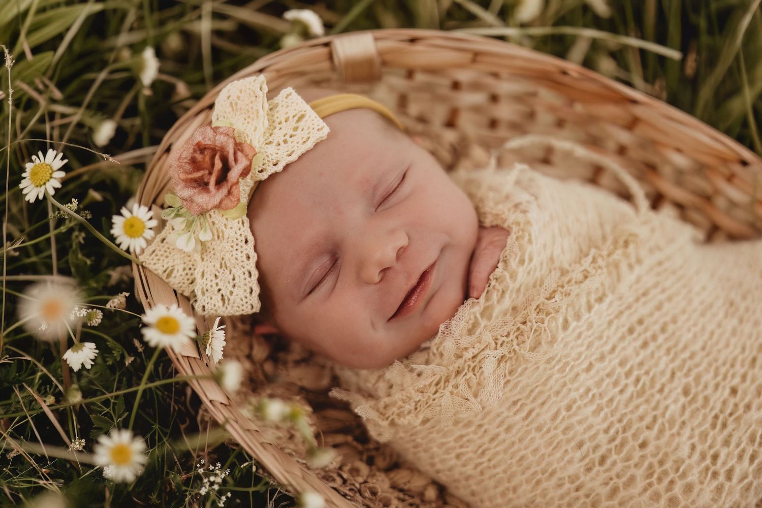 Sleeping newborn baby wrapped in cream knit swaddle with floral headband in wicker basket surrounded by daisies.