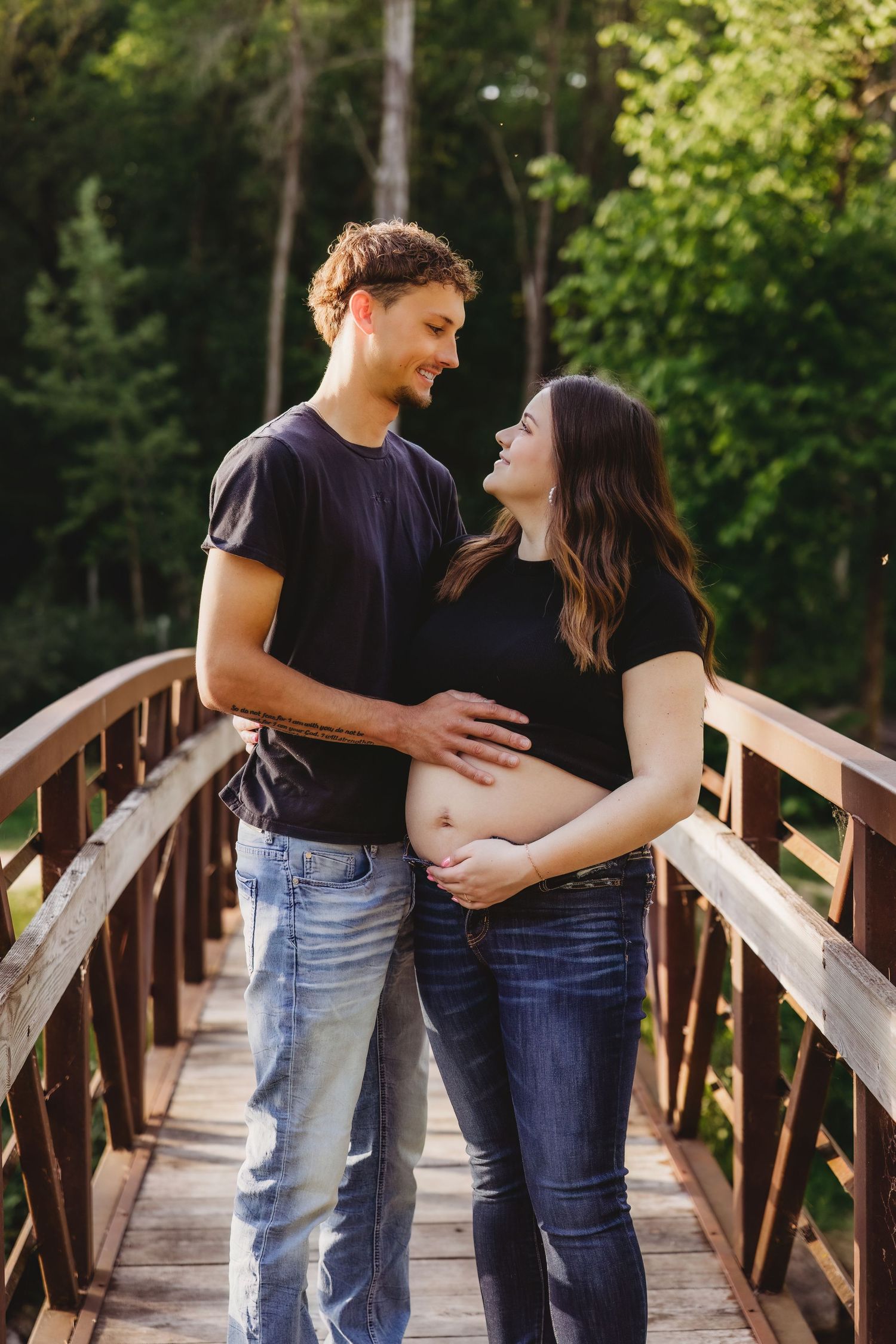 Couple on wooden bridge touching pregnant belly, smiling at each other, surrounded by lush green trees.