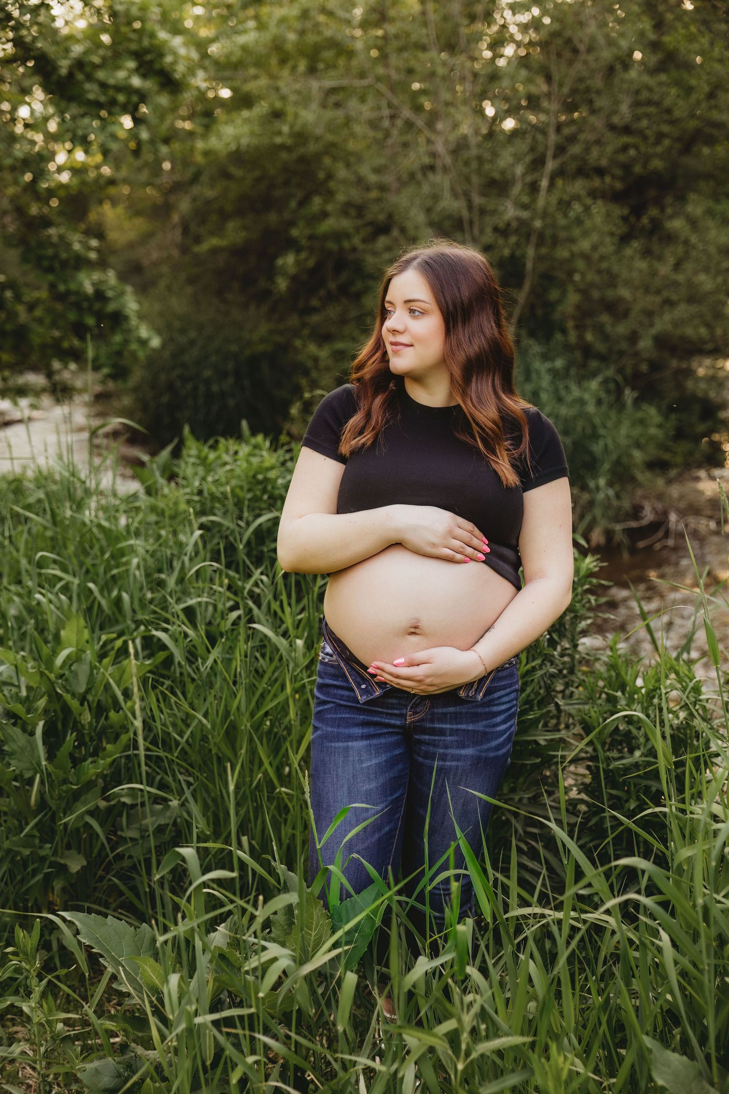 Pregnant woman in black crop top and jeans cradling baby bump outdoors among tall green grass and trees.