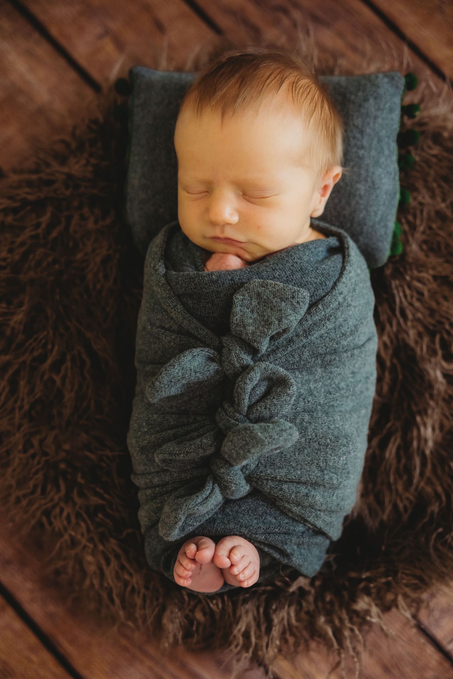 Peaceful newborn baby swaddled in a gray wrap, sleeping on a brown fur blanket over rustic wood flooring.