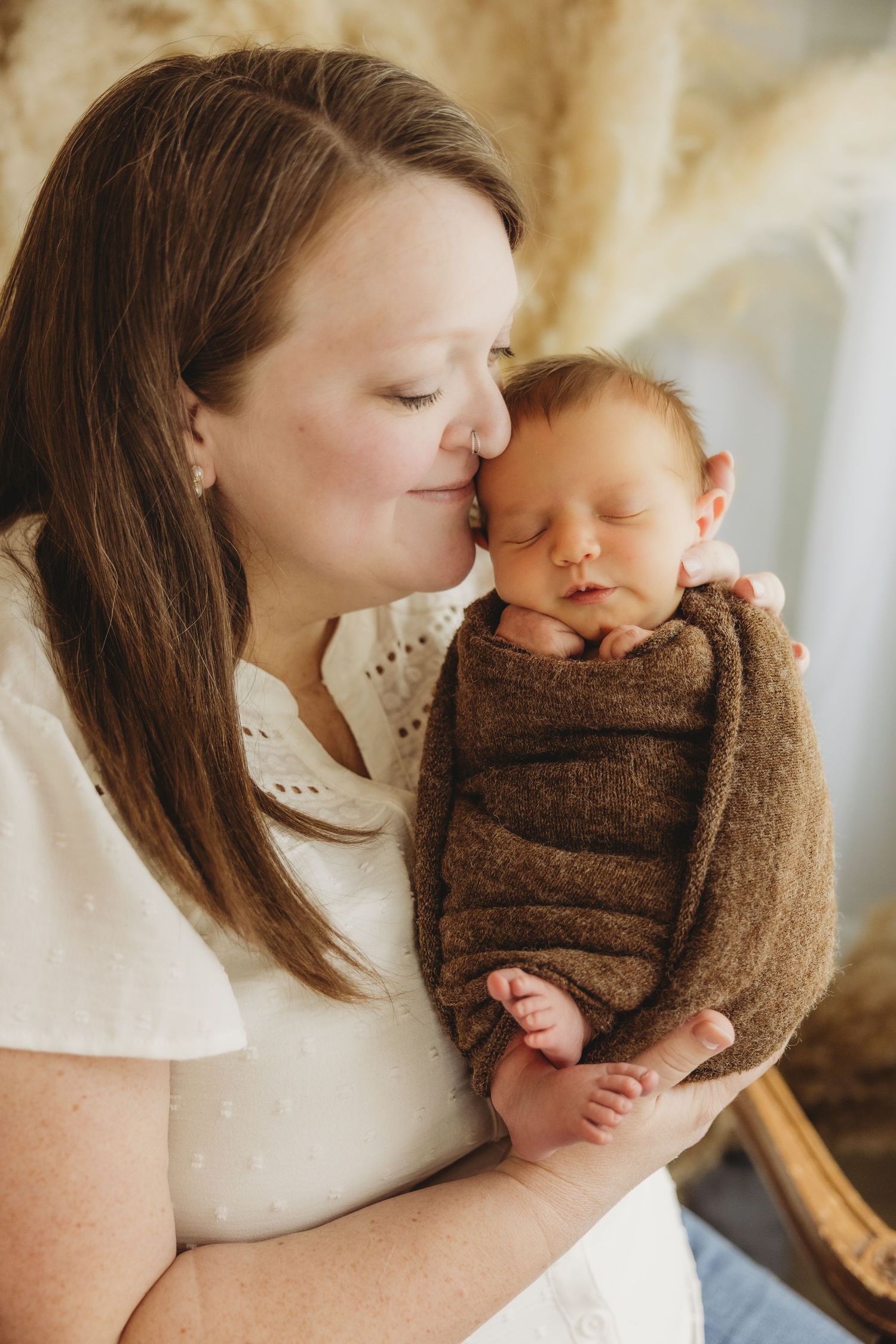 A mother tenderly kisses her sleeping newborn baby wrapped in a brown blanket during a professional newborn photo session.