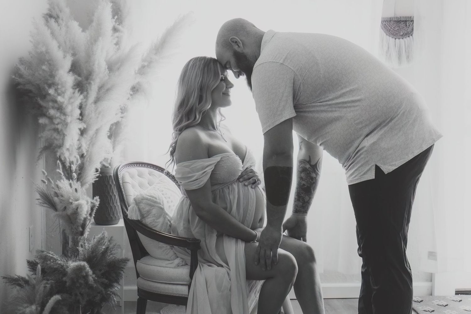 Black and white maternity photo of pregnant woman sitting in chair with partner leaning in for a kiss.