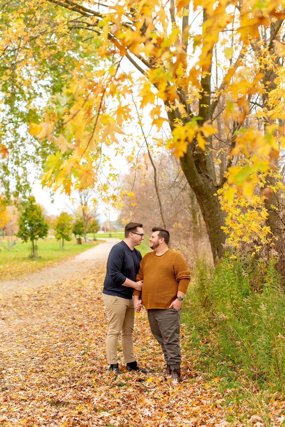 A couple stands together on a leaf-covered path beneath yellow autumn trees in Guelph.