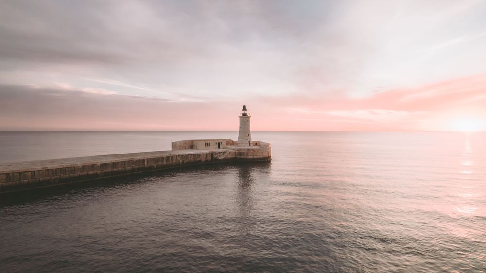 Lighthouse on pier during pink sunset over calm ocean waters.