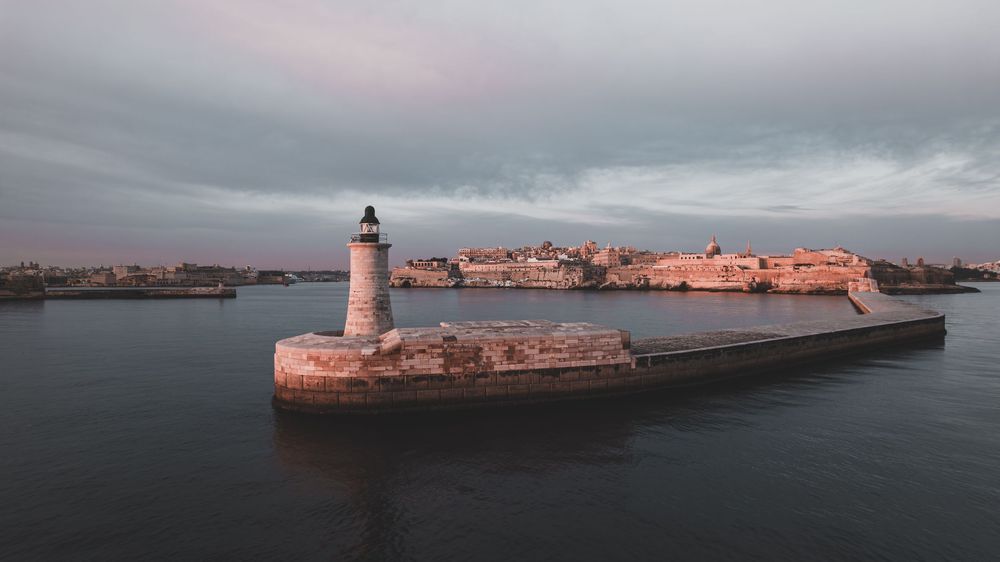 Panoramic view of a lighthouse on a rocky pier during sunset with calm waters and cloudy skies.