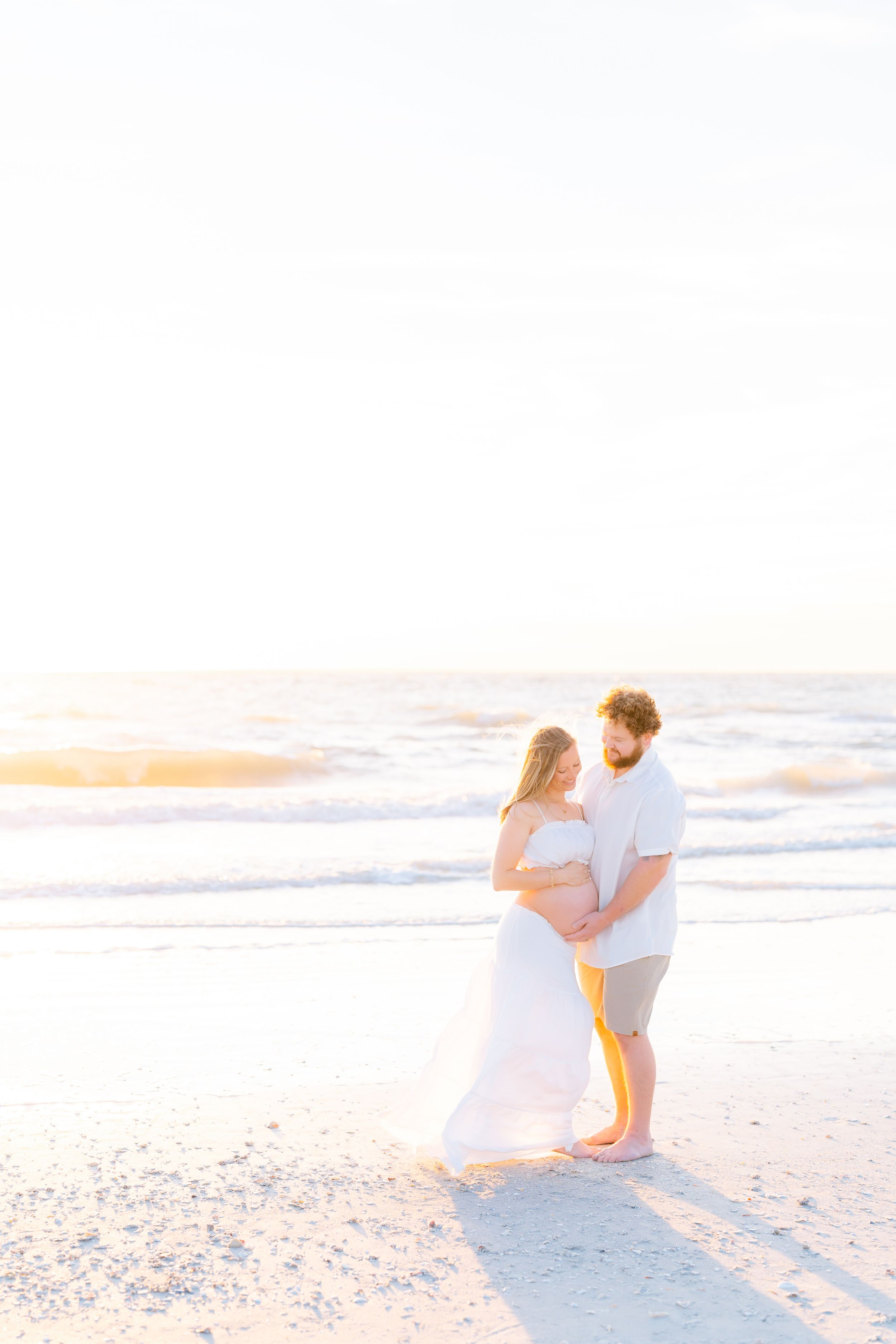 mom and dad looking down at belly during their pregnancy maternity photoshoot on st. Pete beach at golden hour emotional