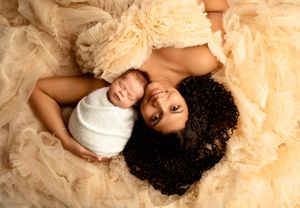 Mother in beige gown lying with swaddled newborn baby on soft tulle fabric.