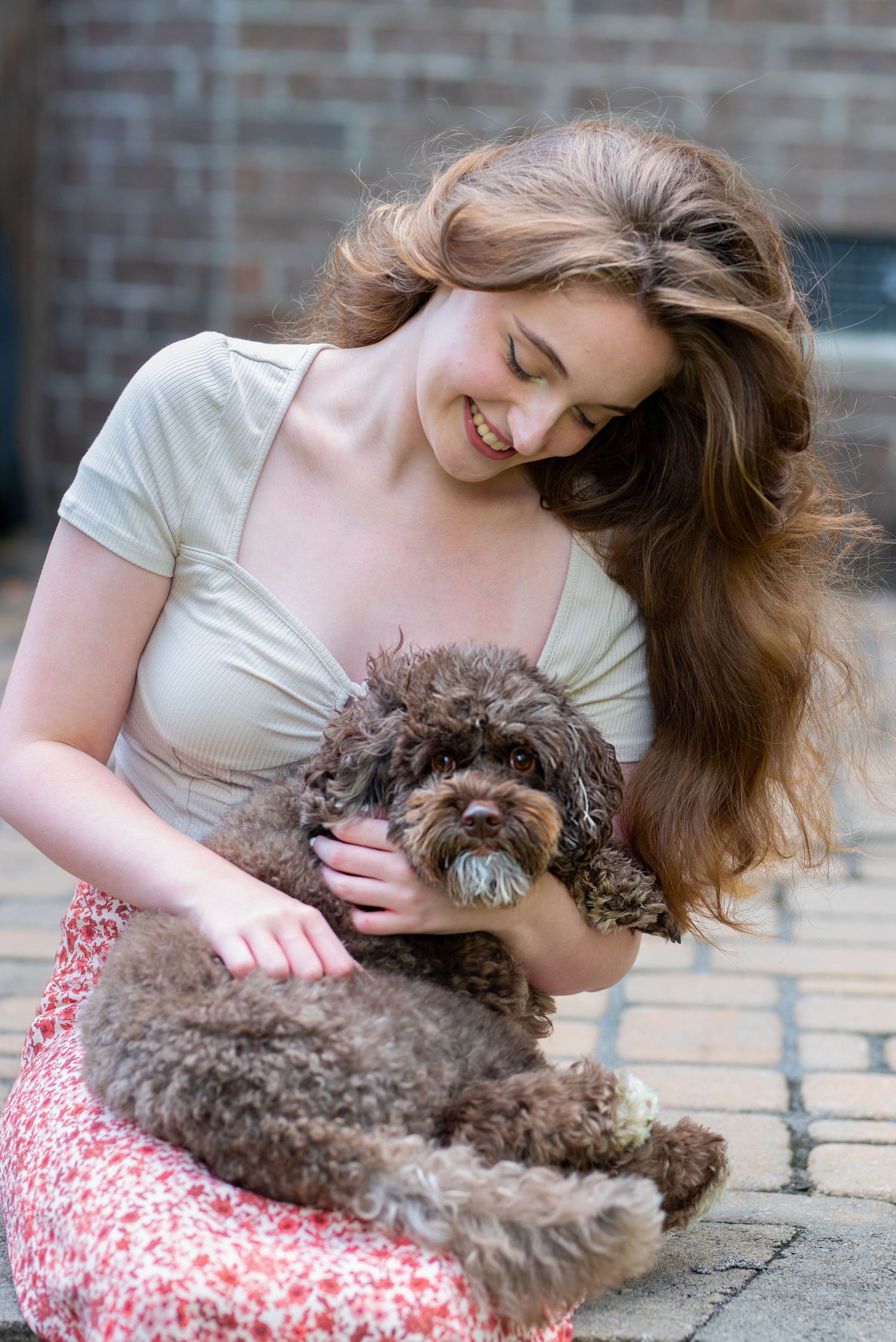 Person with long hair cuddling chocolate brown poodle on brick patio.