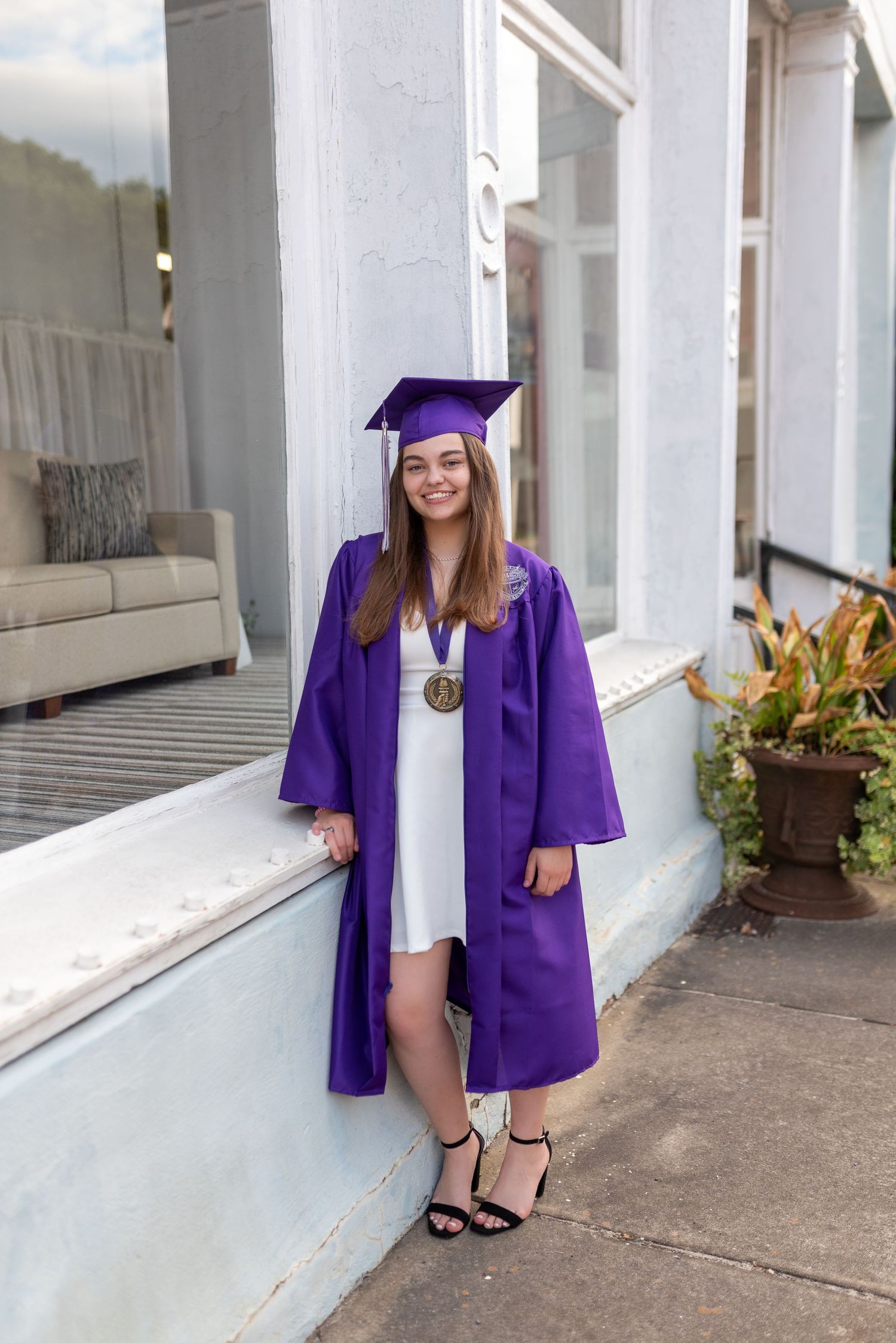 Graduate wearing purple cap and gown poses in front of white building with medal.
