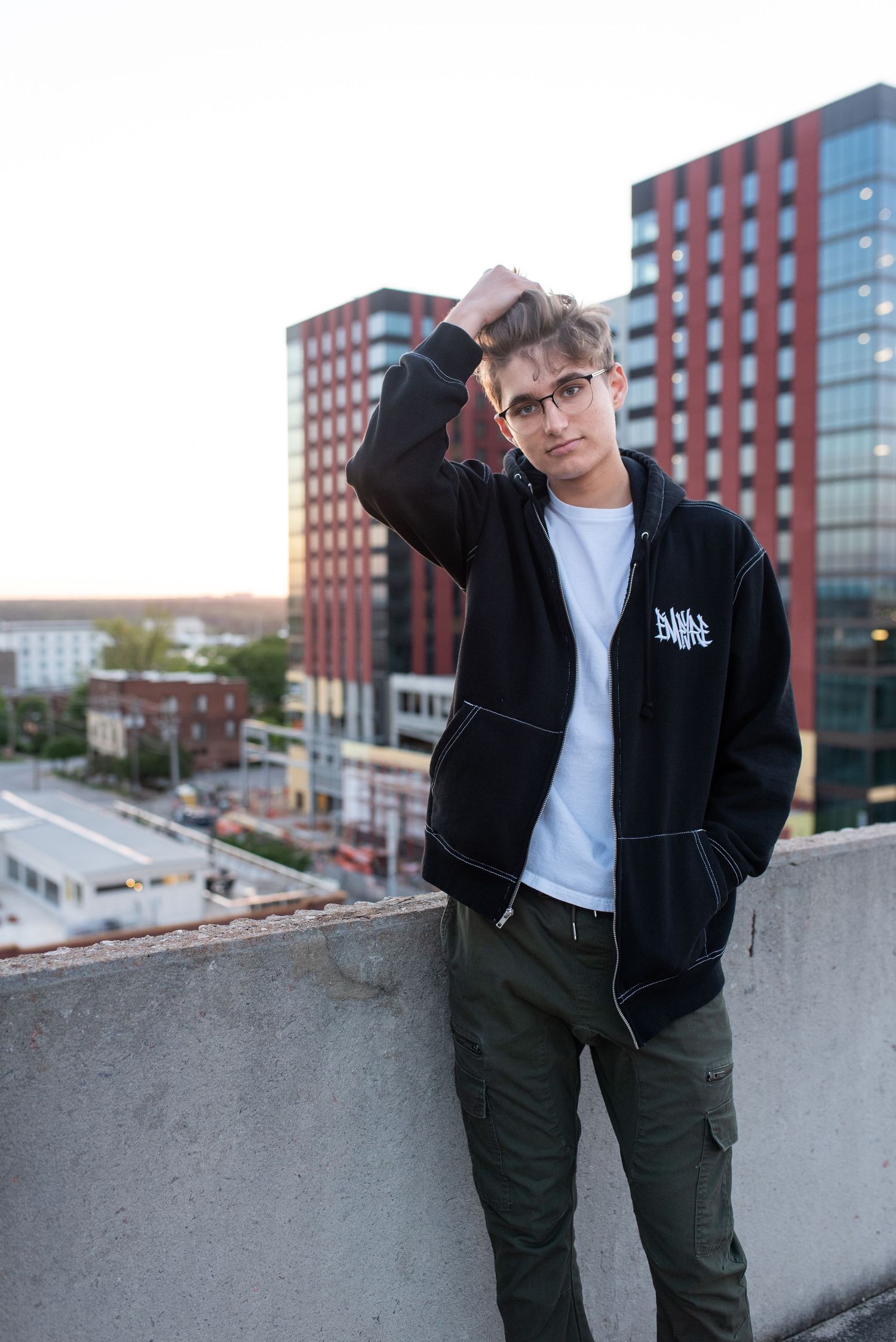 Person in black jacket poses on rooftop with urban buildings in background.
