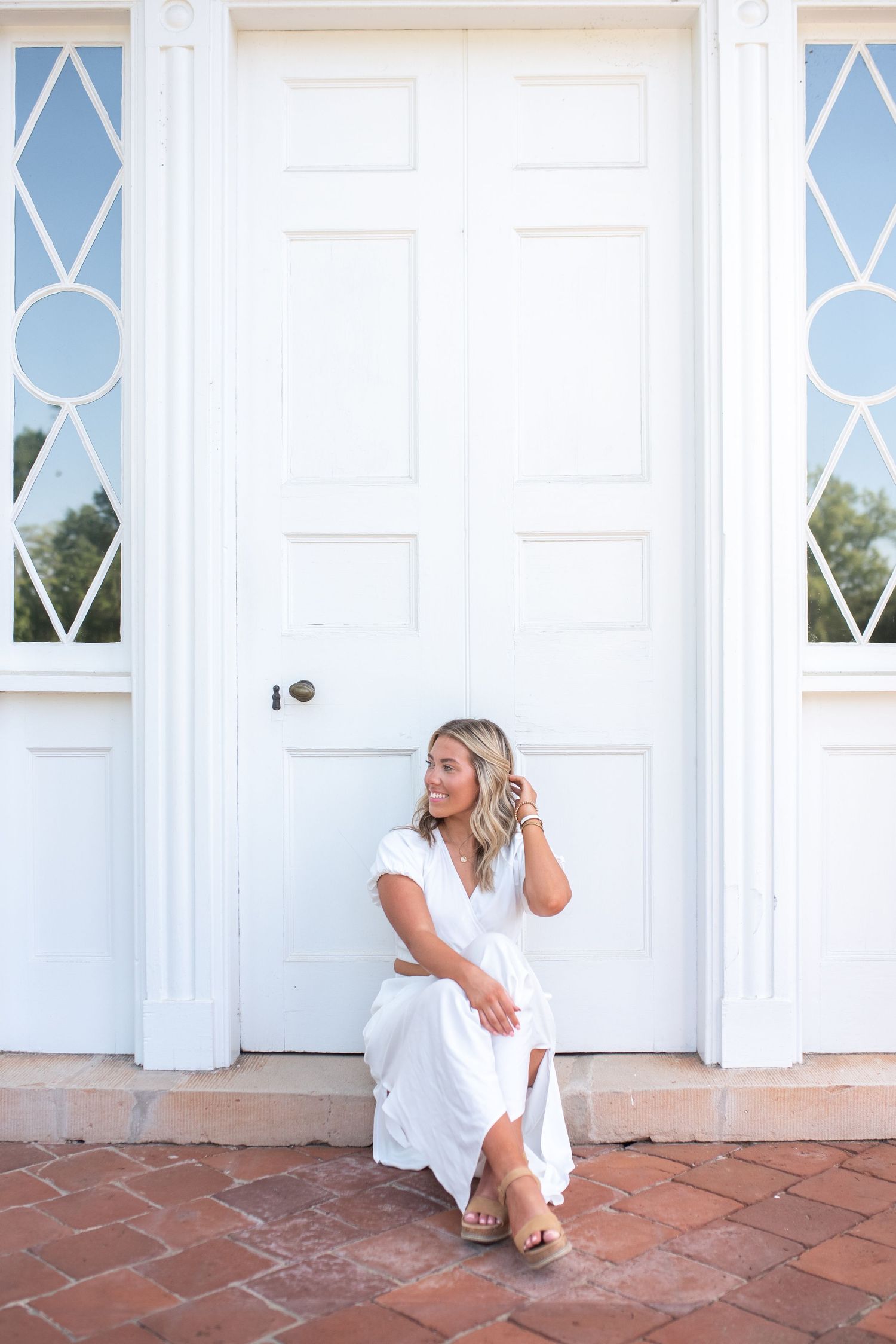 Person in white dress poses on red brick porch with white colonial architecture.