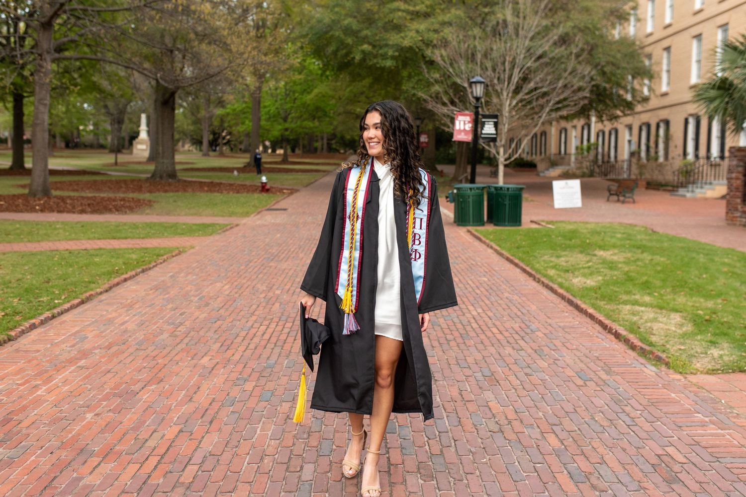 Person walking on brick pathway wearing graduation cap and gown on college campus.