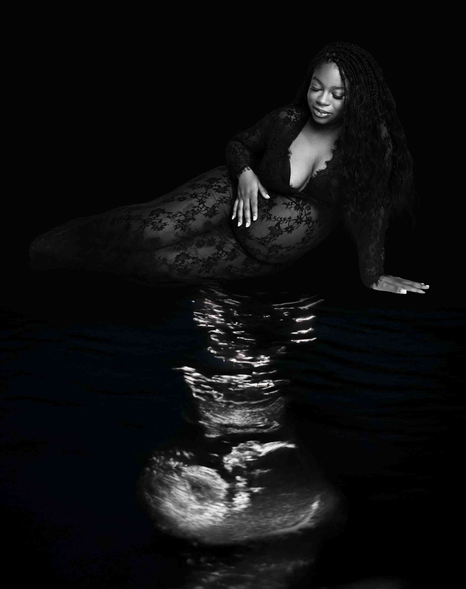 Black and white photo of a woman in lace outfit posing gracefully beside reflective dark water. studio maternity photoshoot cypress texas