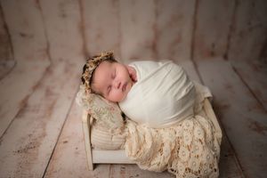Sleeping newborn wrapped in white swaddle with lace blanket and floral crown on rustic wooden backdrop.