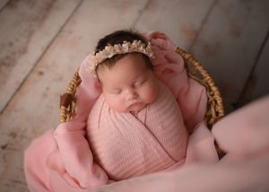 Sleeping newborn wrapped in soft pink swaddle with floral crown in woven basket.