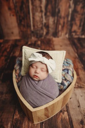 Peaceful sleeping newborn wrapped in gray swaddle in heart-shaped wooden basket.