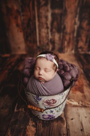 Newborn wrapped in gray swaddle sleeping peacefully in vintage floral teacup on wooden surface.