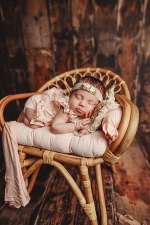 Peaceful newborn in lace outfit sleeping on vintage rattan chair against rustic backdrop.