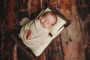 Swaddled newborn sleeping in rustic wooden box on dark wooden floor.