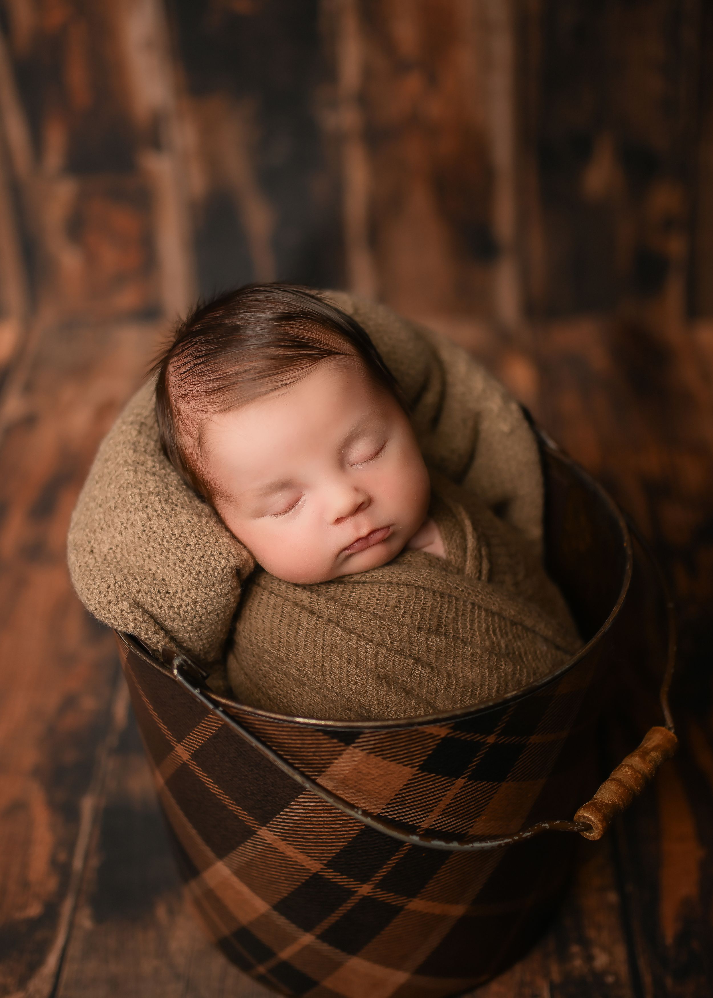 Sleeping newborn wrapped in brown knit fabric nestled in a plaid basket against dark wood background.