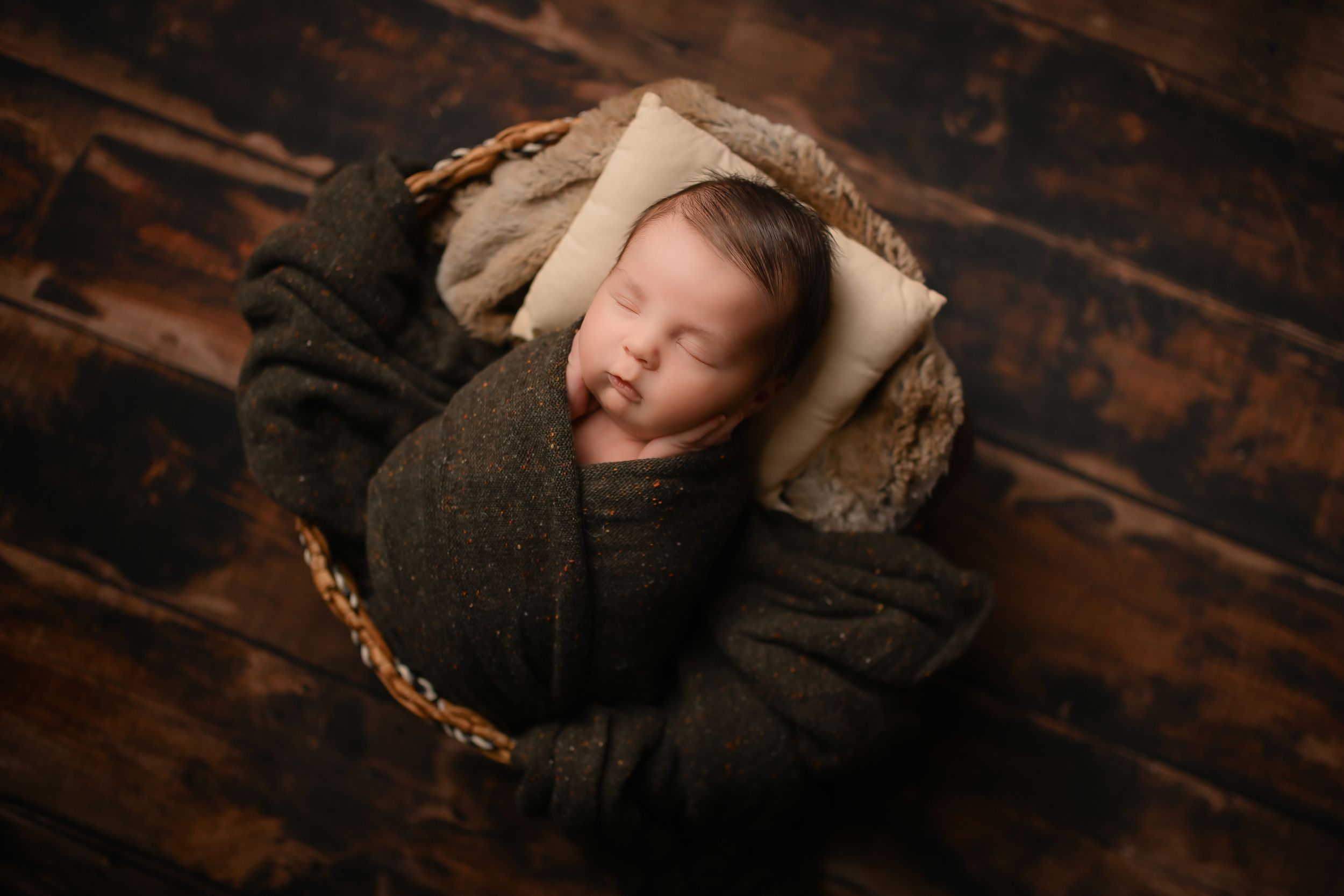 Newborn baby sleeping peacefully in dark wrap inside rustic wooden basket.