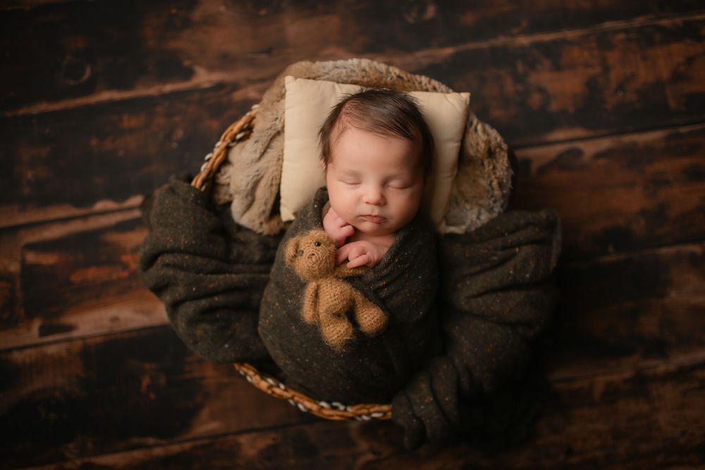 Sleeping baby wrapped in dark fabric lying in round basket with stuffed toy.