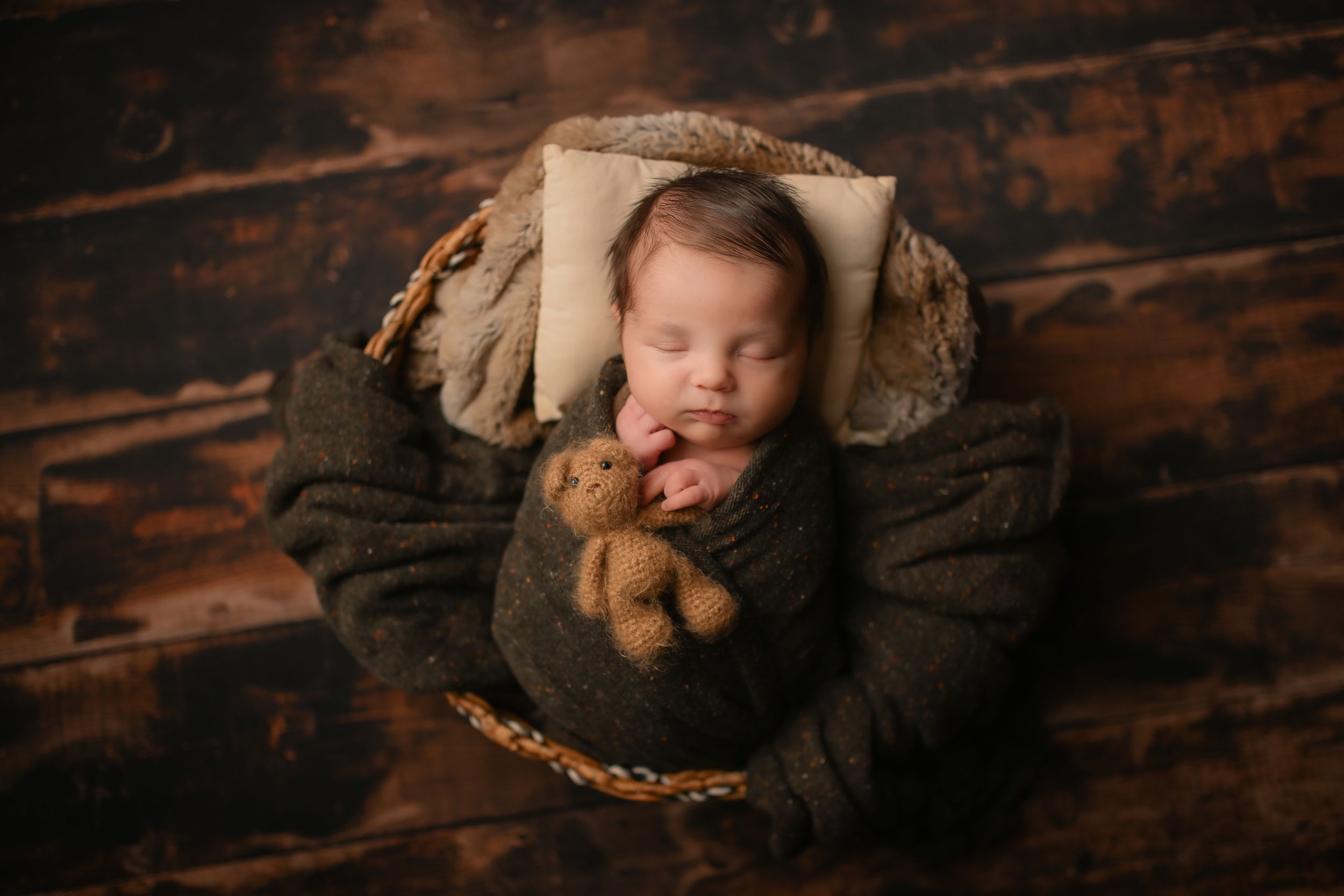 Sleeping baby wrapped in dark fabric lying in round basket with stuffed toy.