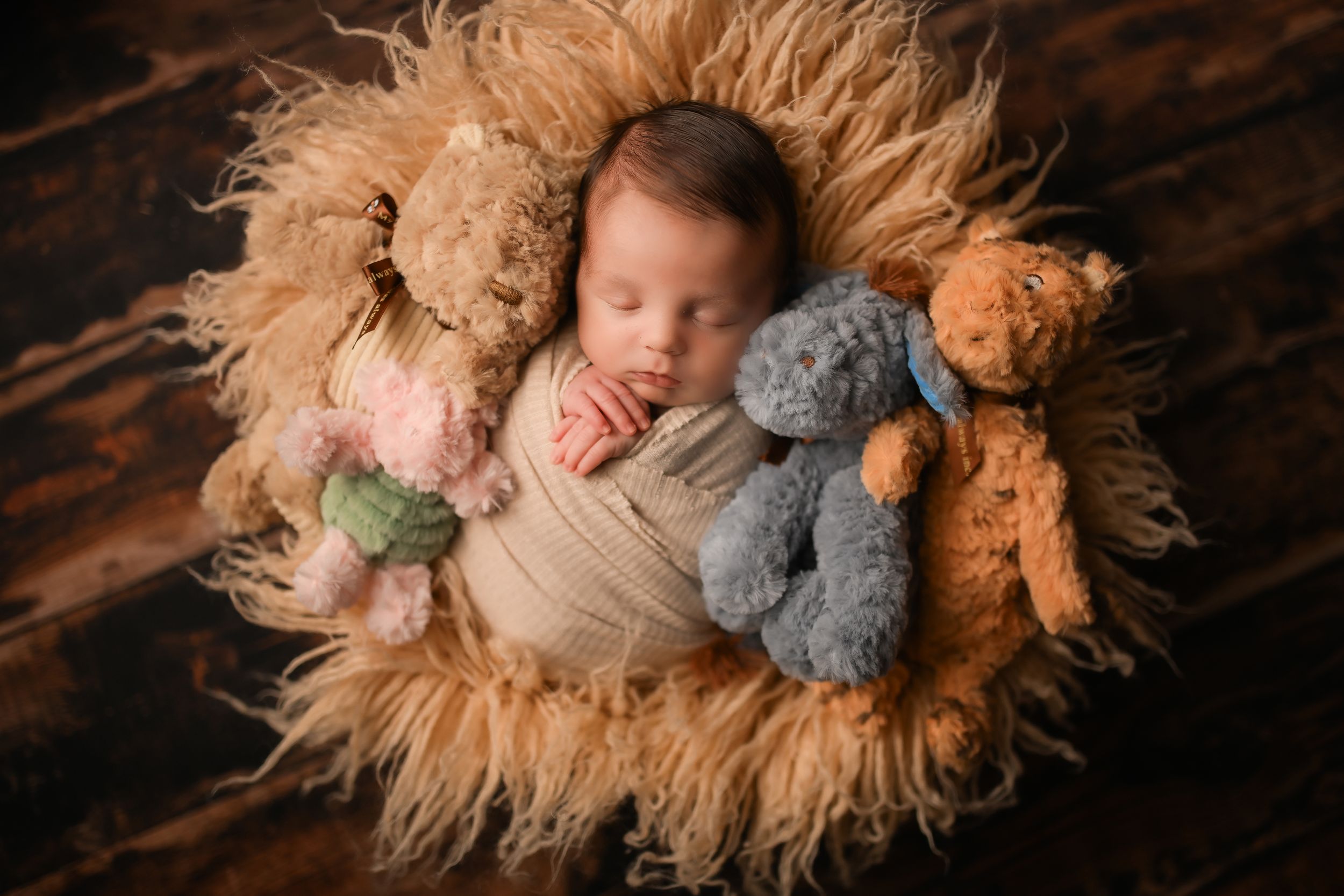 Sleeping newborn on furry blanket surrounded by plush teddy bears against wooden background.