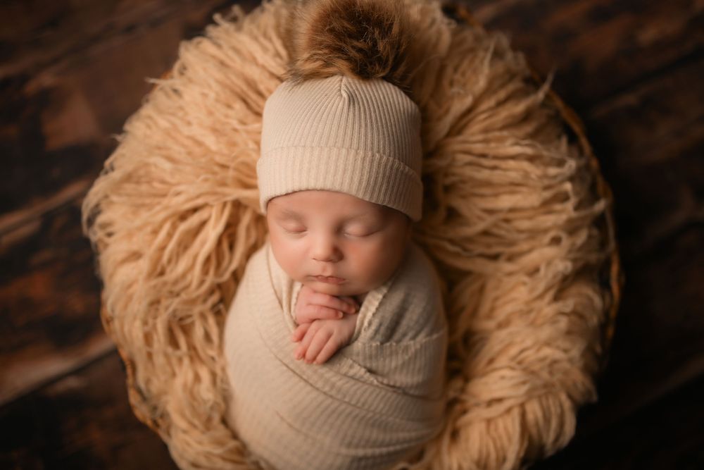 Sleeping baby in beige hat and wrap nestled in furry tan nest.