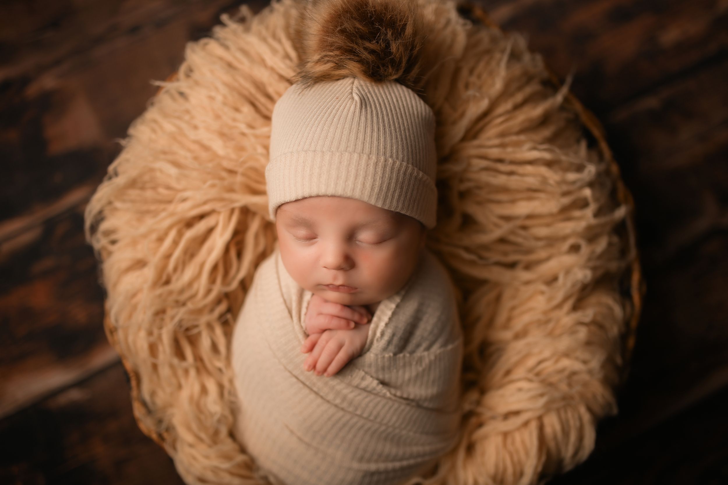 Sleeping baby in beige hat and wrap nestled in furry tan nest.