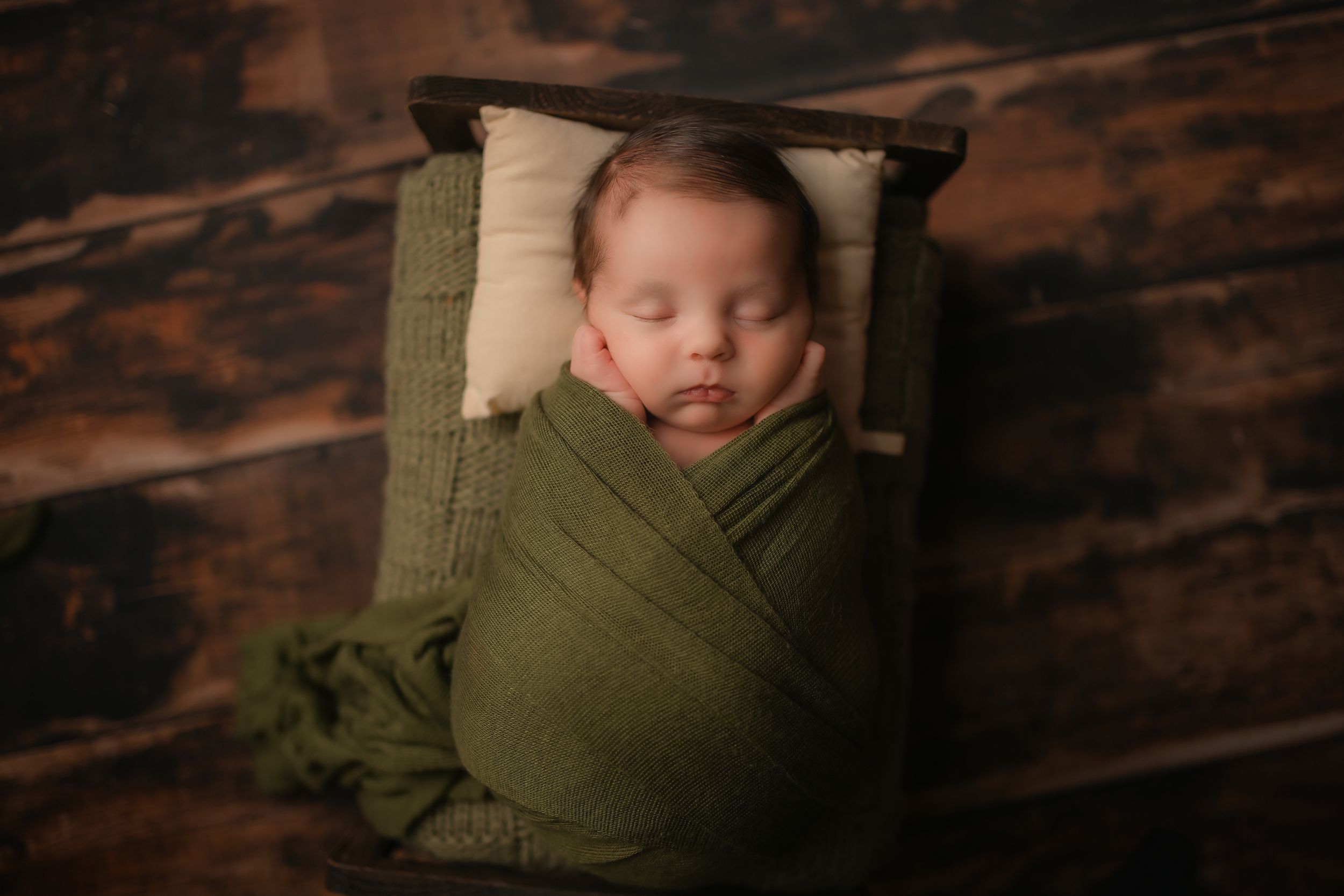 Sleeping newborn wrapped in olive green swaddle on small white pillow.