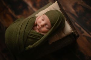 Sleeping baby swaddled in dark green wrap resting on white pillow.
