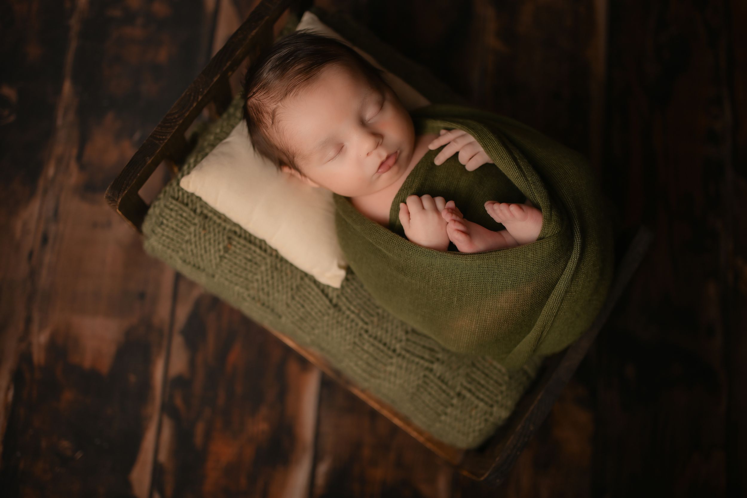 Newborn sleeping in olive green wrap on cushion against dark wooden background.
