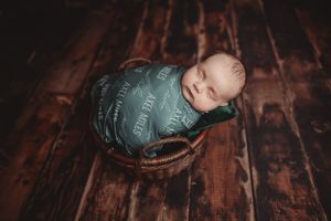 Sleeping newborn wrapped in teal swaddle nestled in vintage basket on wooden floor.