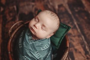 Close-up of sleeping newborn in green patterned wrap against dark wooden background.