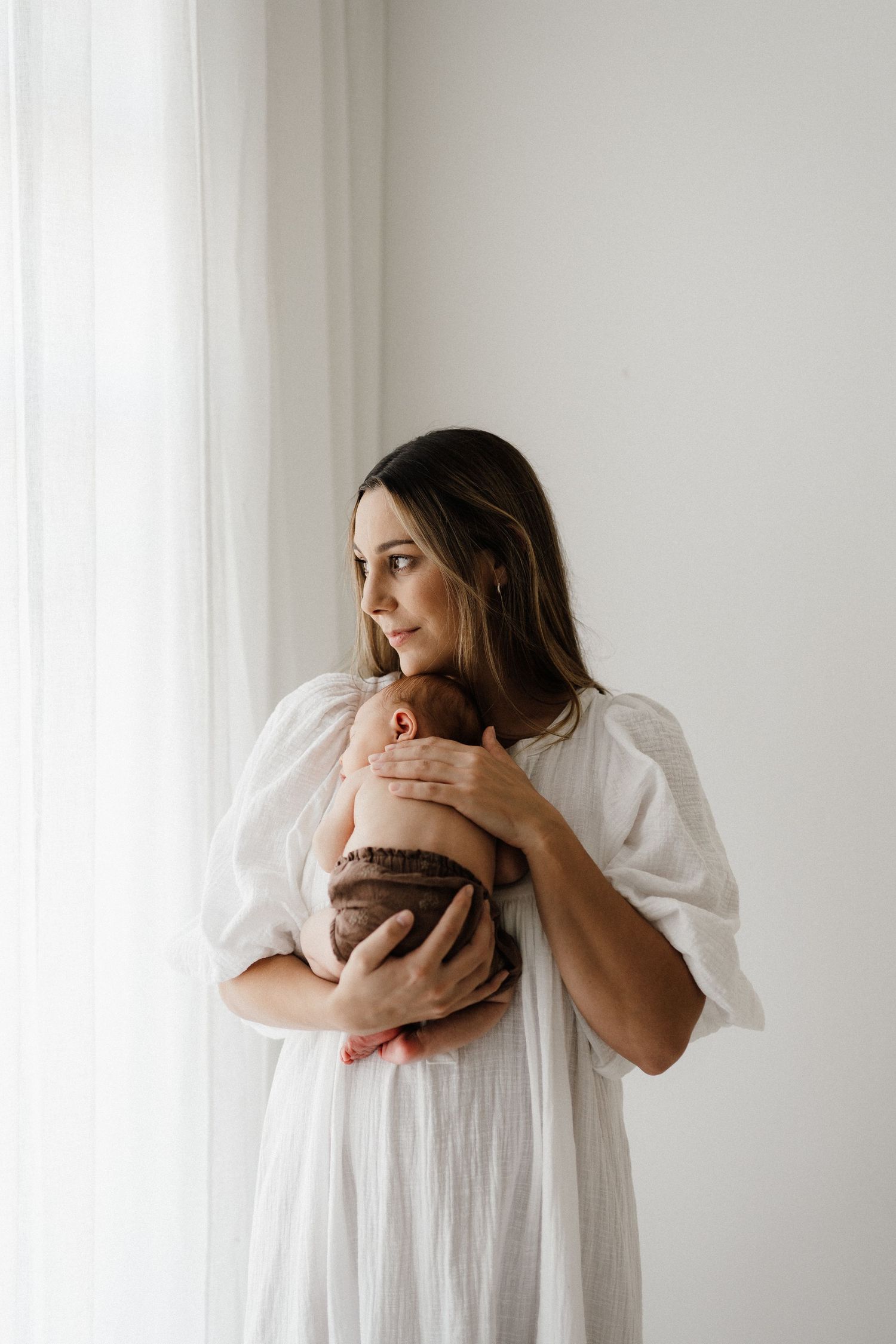Woman in white dress holding baby near window in minimalist setting.