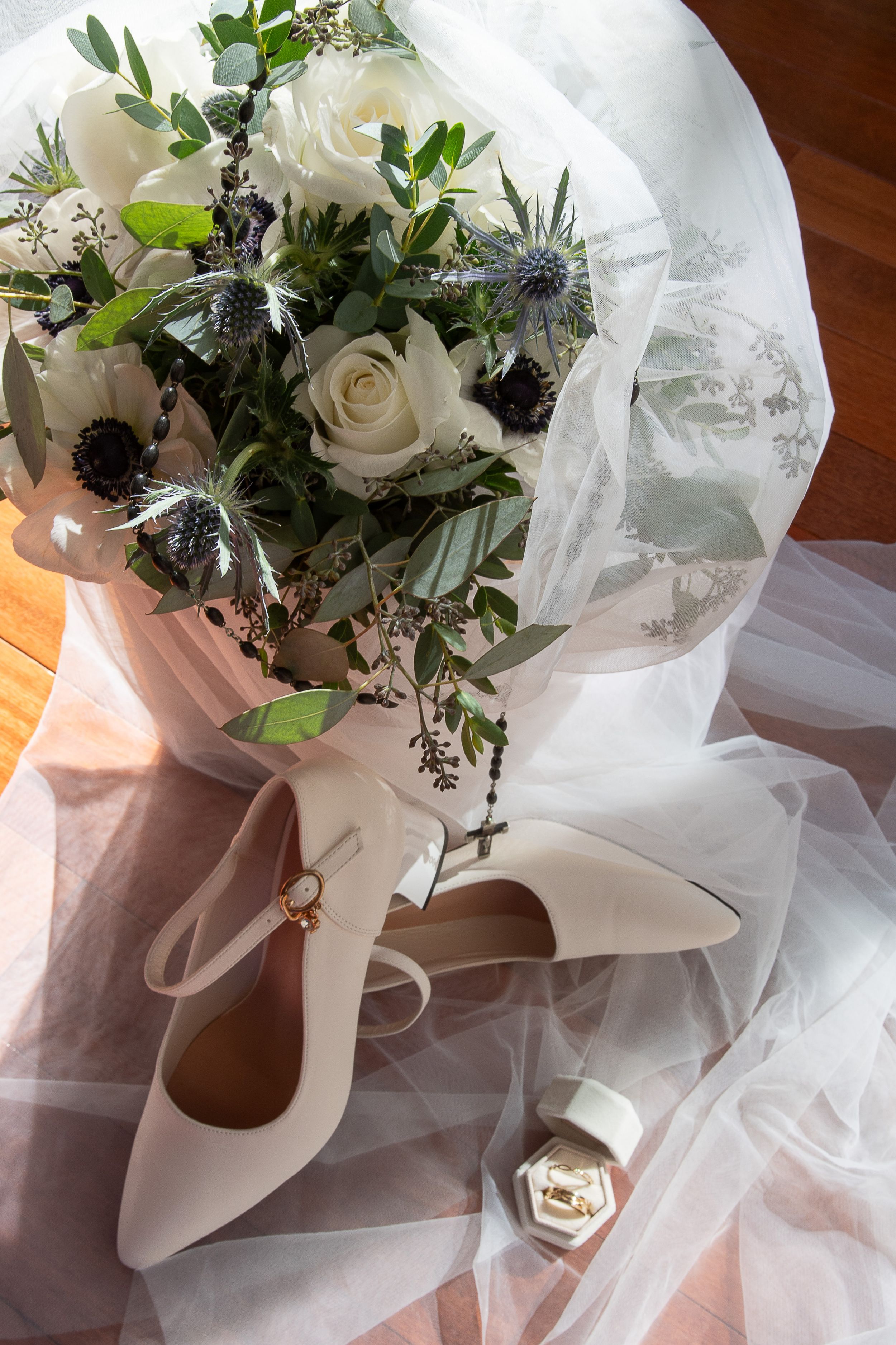 Wedding bouquet with white roses and blue thistle wrapped in delicate tulle and satin ribbon rests on wooden surface.