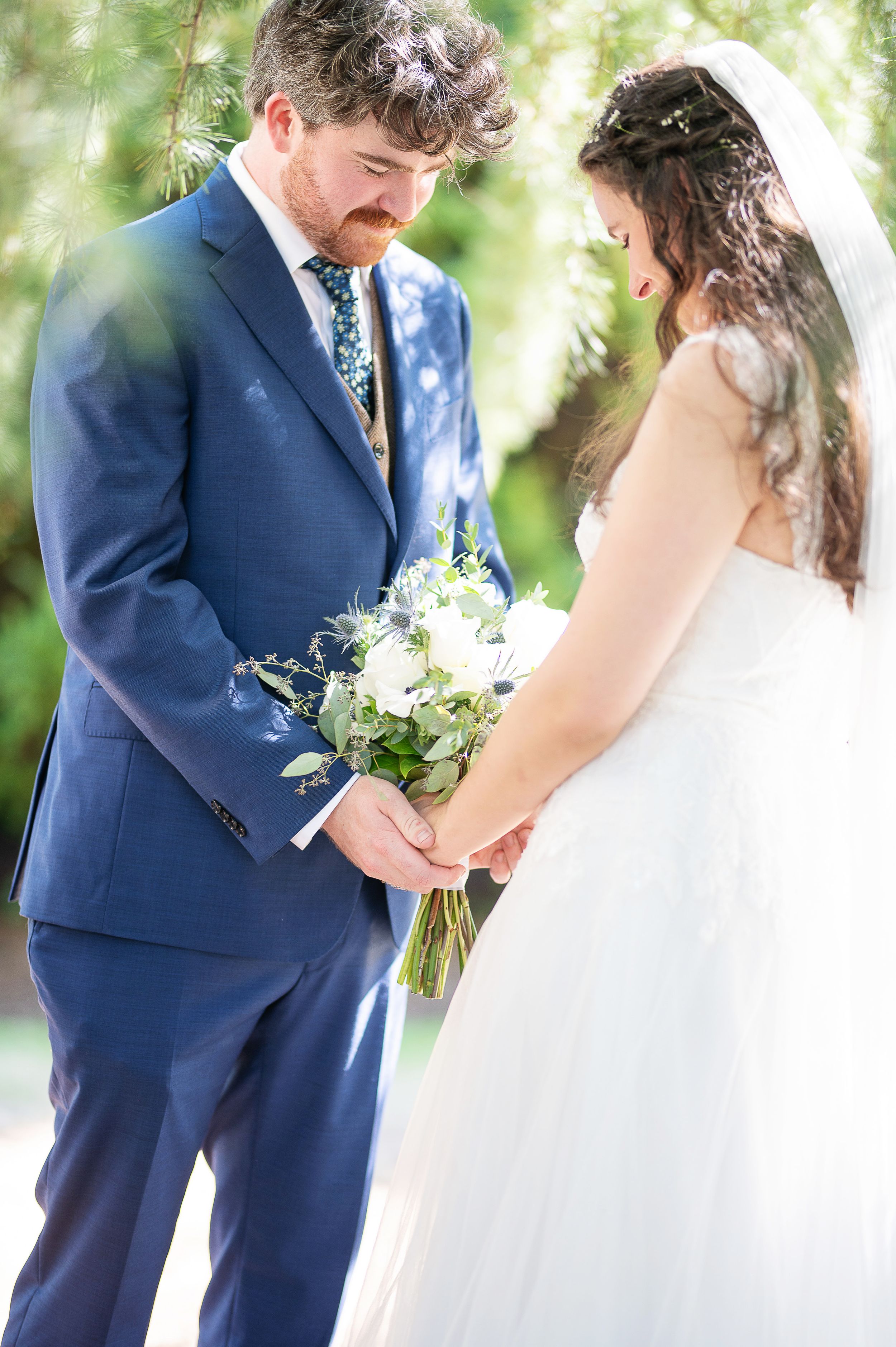 A romantic wedding moment captured in a garden setting with a couple in navy suit and white dress holding a bouquet.