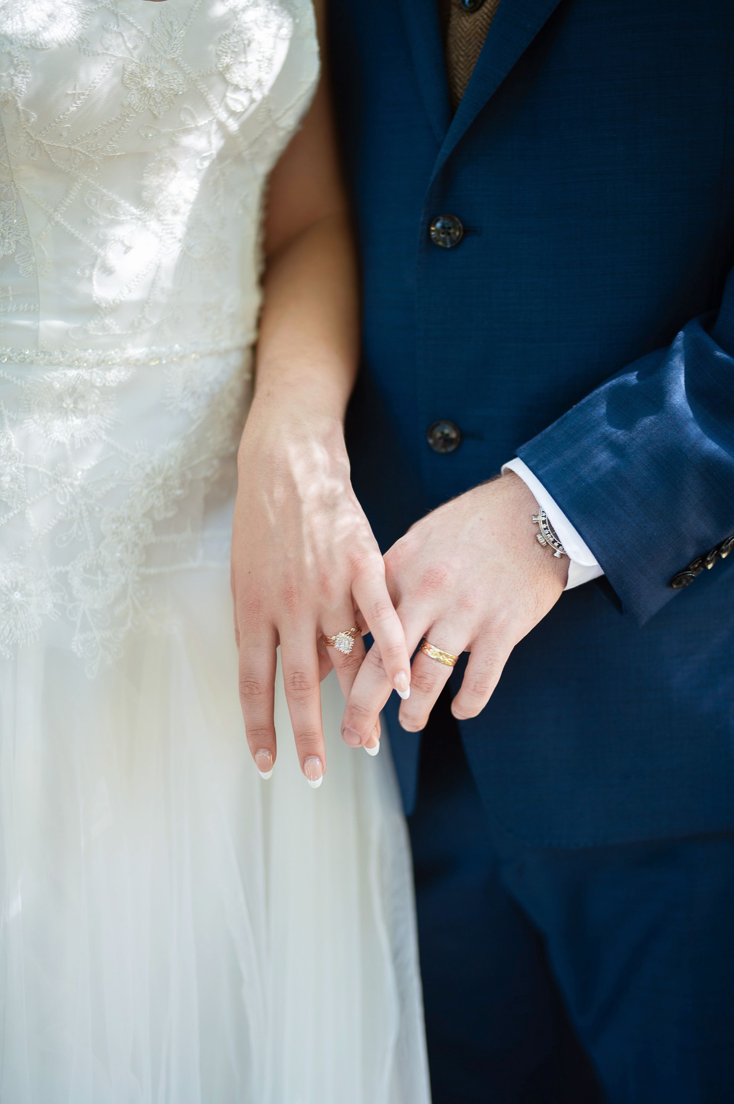 Close-up of newlyweds' hands with wedding rings against white dress and navy blue suit.