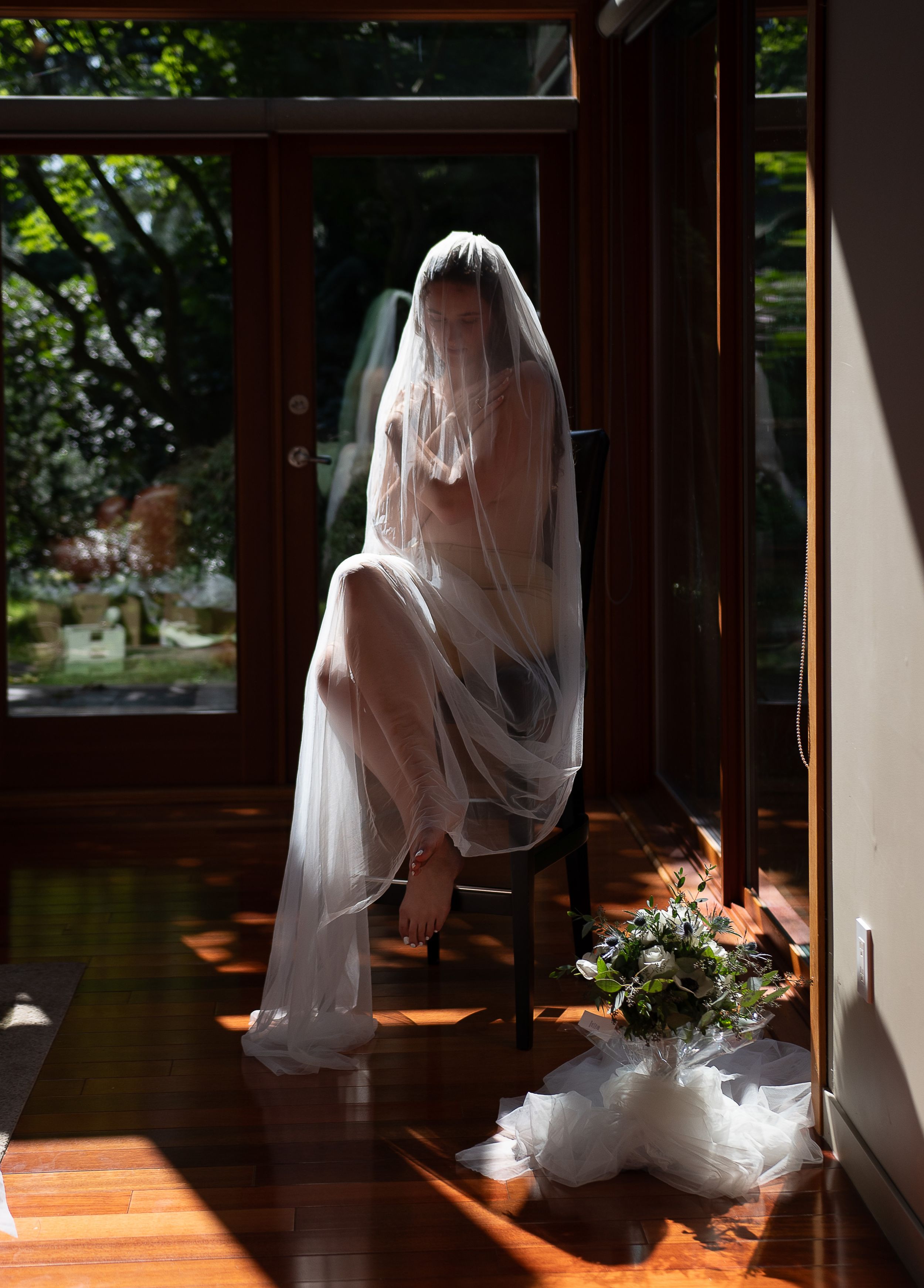 A bride in a flowing white dress and veil stands silhouetted against sunlit windows in a rustic wooden interior.