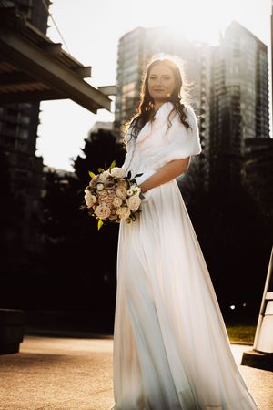 A bride in a flowing white dress holds a bouquet against a cityscape backdrop during golden hour.